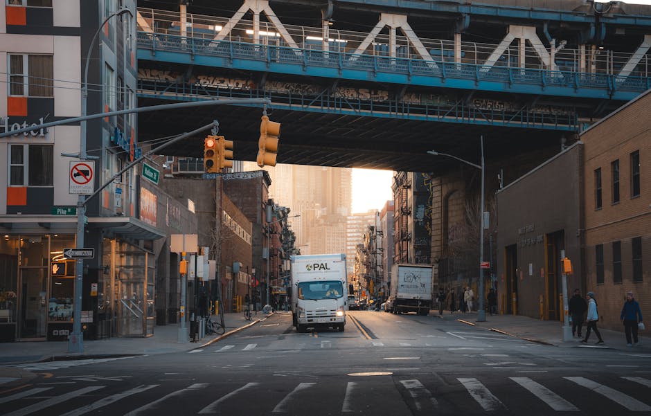A city street scene captured during daylight hours, showing a white merchandise delivery van from PAC positioned centrally on the road beneath a blue elevated railway track or bridge structure. The st