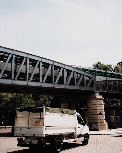 A city street scene captured during daylight hours, showing a white merchandise delivery van from PAC positioned centrally on the road beneath a blue elevated railway track or bridge structure. The st